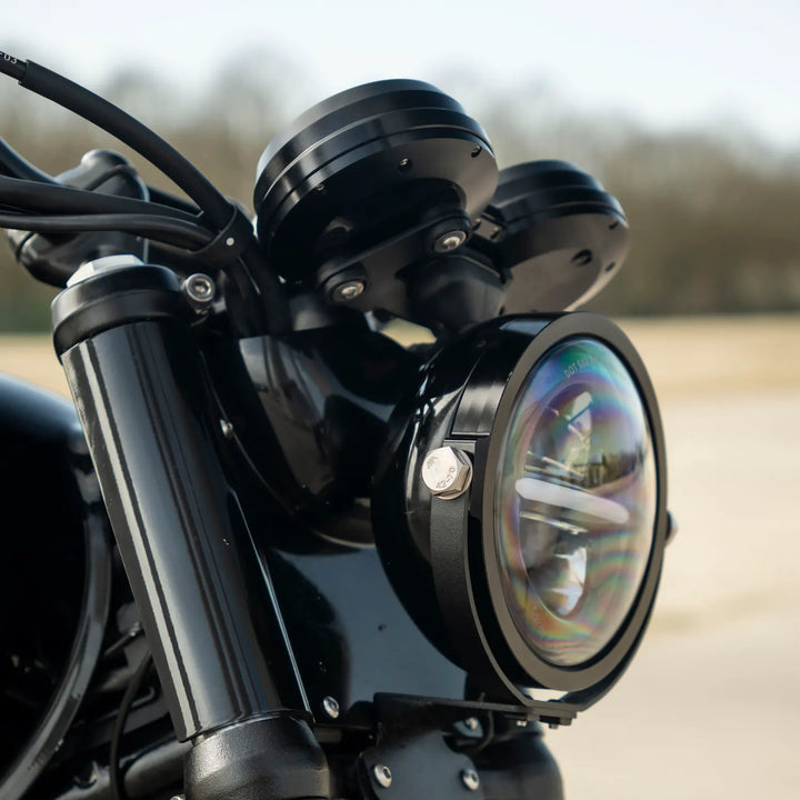 Close-up of a black aluminium bracket fitted around a round LED motorcycle headlight, showing the silver side mounting bolt.