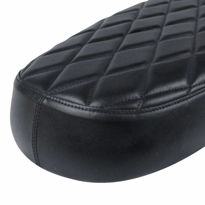 Close-up of a black low-profile motorcycle seat with a textured synthetic leather cover, diamond-quilted top, piping and stitched sides.